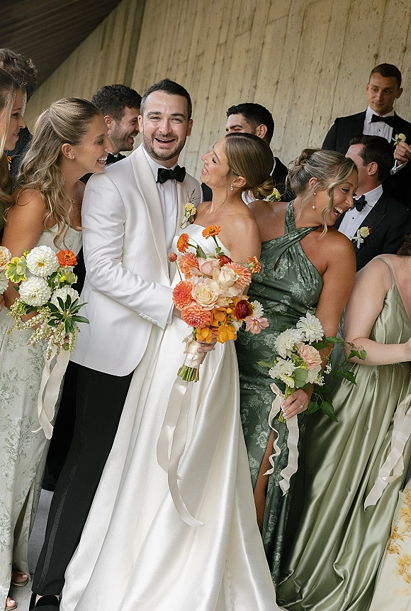 Wedding party photo of bride and groom with bridesmaids laughing with bouquets and satin ribbons under a covered walkway by a wood wall