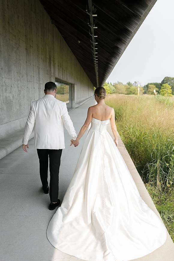 Couple portrait of bride and groom walking away holding hands, her strapless gown train flowing under a modern concrete walkway
