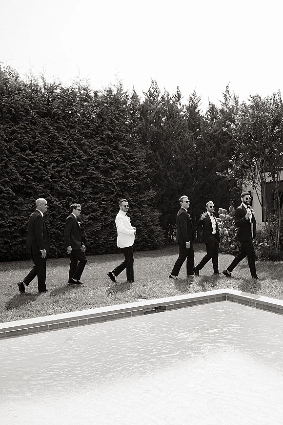 Groomsmen portrait with groomsmen walking photo in tuxedos, bow ties and sunglasses, groom in white jacket by a poolside lawn hedge