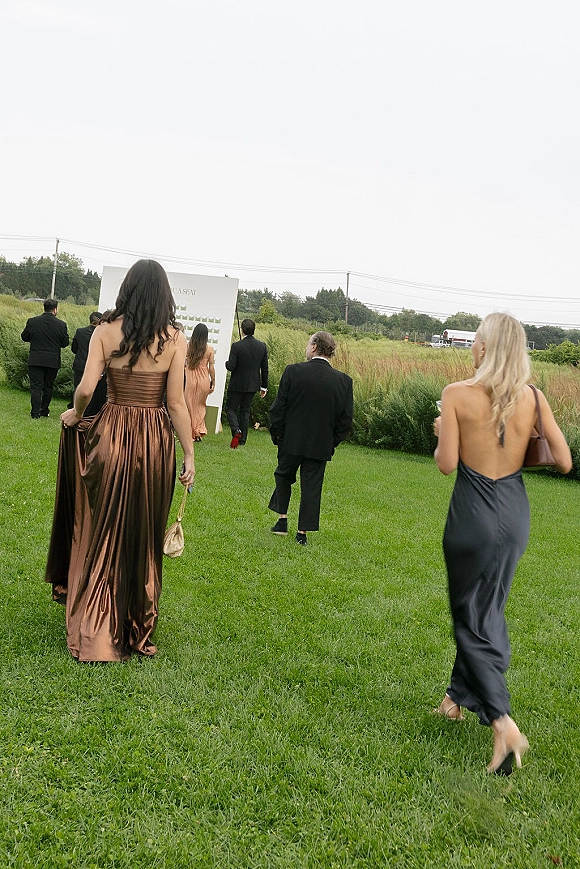 Wedding guest moment as women in evening gowns and men in suits walk across a meadow field under an overcast sky, clutch purses in hand