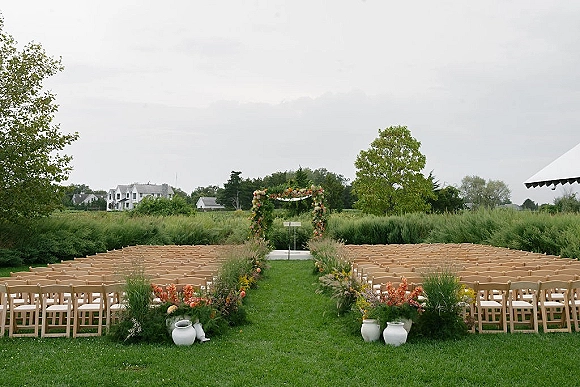 Ceremony setup with outdoor ceremony seating facing a floral arch, wood folding chairs and aisle flowers on a meadow lawn under clouds