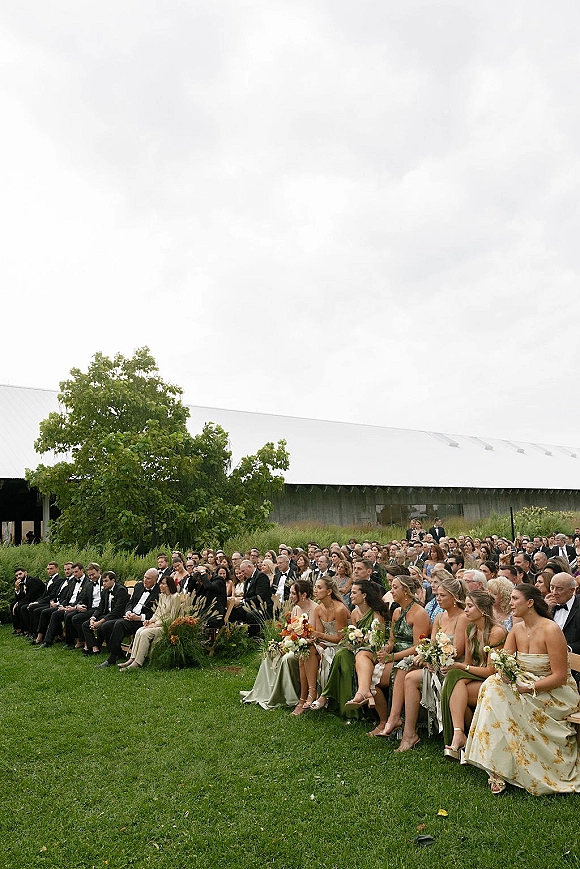 Ceremony guests seated in outdoor wedding ceremony seating, with bridesmaid bouquets and tuxedos with black bow ties on a grassy lawn near a barn