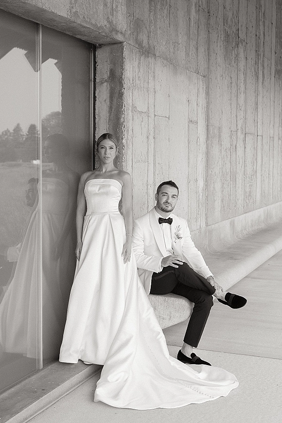 Couple portrait in a black and white wedding portrait style, bride in strapless gown with long train beside groom in white jacket by concrete wall window