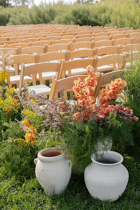 Ceremony aisle decor with wood folding chairs and white cushions, flanked by clay pots of orange orchids and yellow blooms on grass lawn