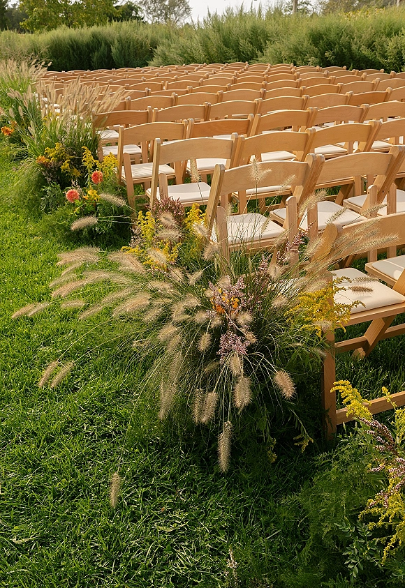 Ceremony seating with outdoor ceremony chairs in neat rows, tan wood folding chairs with cushions beside wildflower aisle florals on a lawn