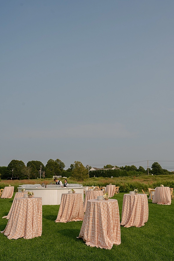Outdoor cocktail hour with wedding cocktail hour setup of patterned high-top tables, floral bud vases and glassware on a lawn by trees and houses