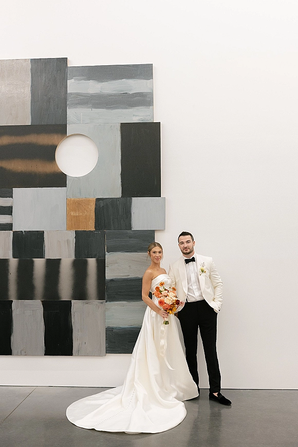 Couple portrait of bride in strapless dress holding a bouquet beside groom in white tuxedo, posed by a modern art wall in gallery setting