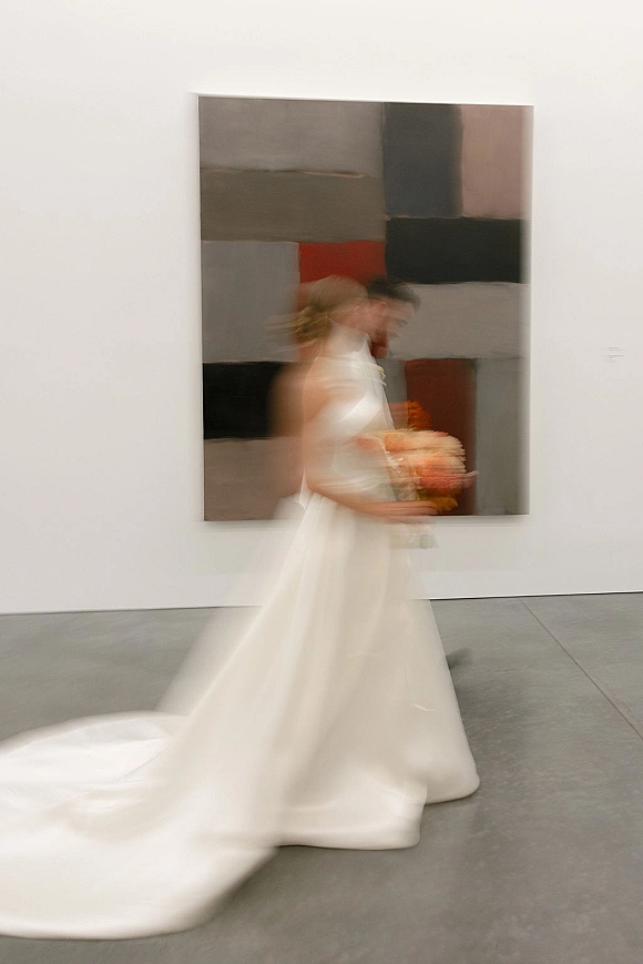 Bridal portrait of a bride walking with bouquet in a white halter wedding dress, motion blur in an art gallery with abstract painting backdrop
