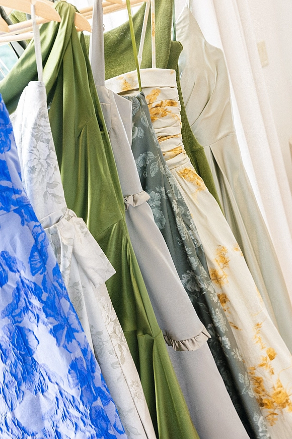 Bridesmaid dresses in a mix and match bridesmaid dresses palette hang on a rack in window light, showing satin and floral prints on hangers