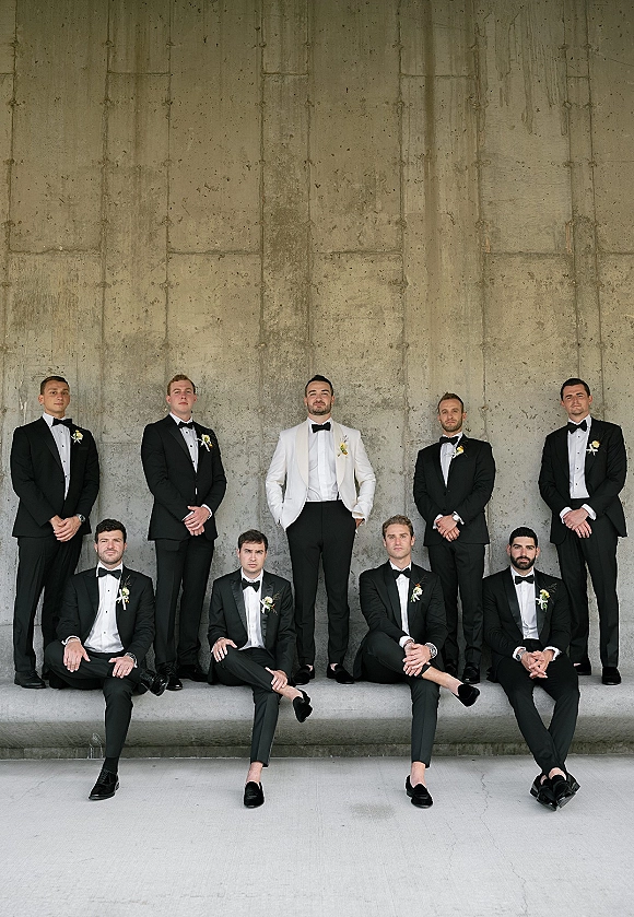 Groomsmen portrait with groom in a white dinner jacket and bow tie among tuxedos and boutonnières against a concrete wall backdrop