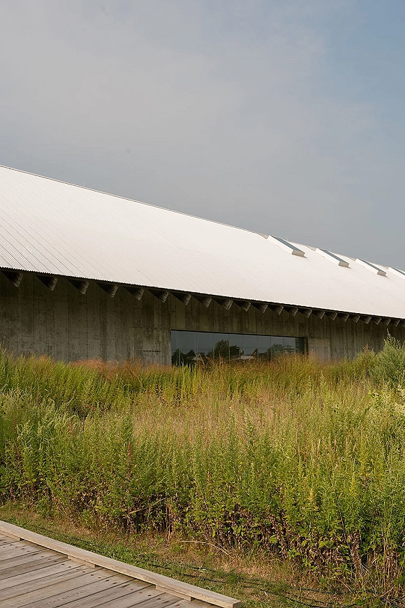 Modern wedding venue with a metal roof and concrete walls, glass windows facing tall grass and a boardwalk path under open sky