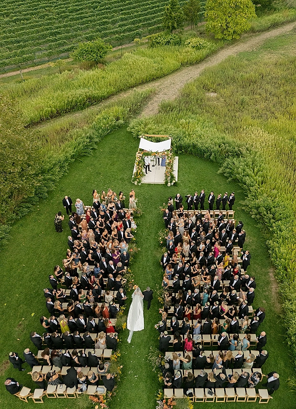 Wedding ceremony outdoors, aerial view of bride walking down a floral-lined aisle to a draped arch on a lawn by vineyard rows