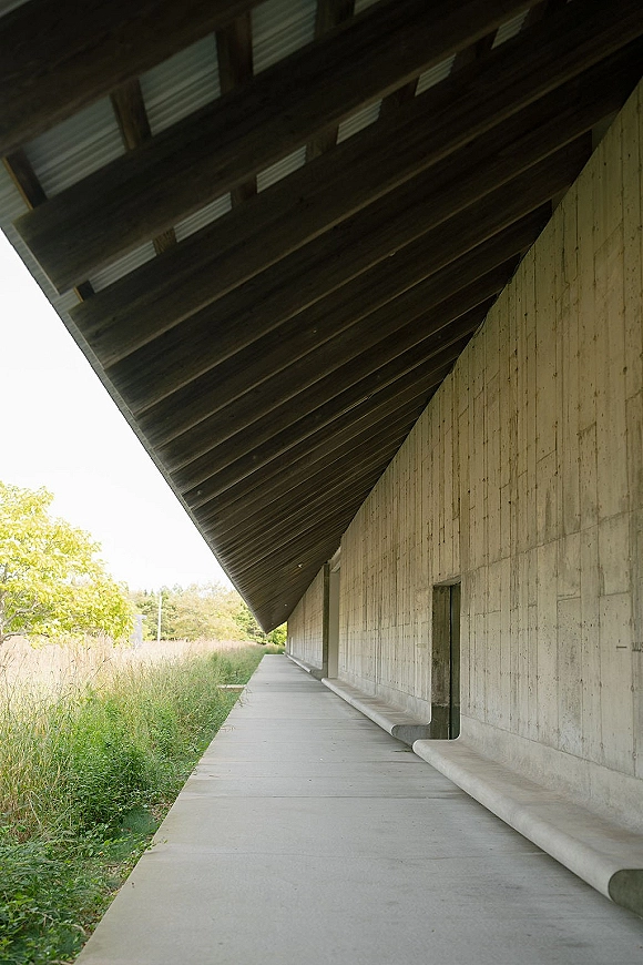 Modern venue exterior with a brutalist wedding venue concrete wall and covered walkway under an overhanging roof, bordered by tall grass and trees