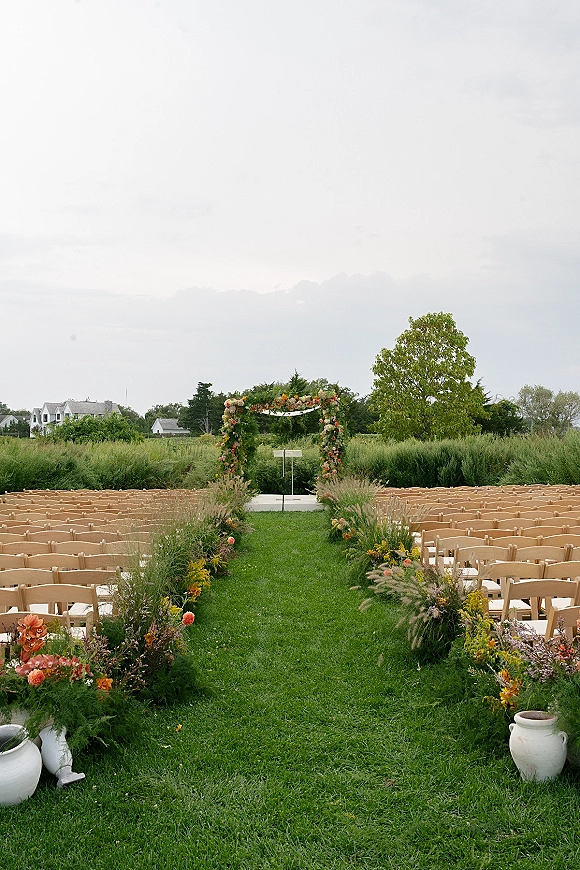 Ceremony setup with outdoor wedding ceremony aisle flowers, asymmetrical floral arch and wildflower urns on a grassy meadow under cloudy sky