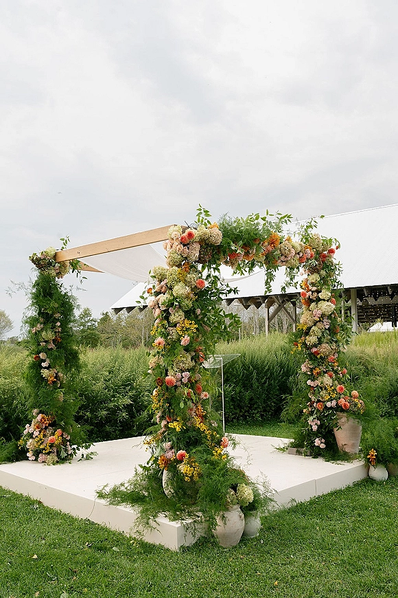 Wedding ceremony arch with a floral ceremony arch of mixed blooms and greenery on a white platform, set on a lawn near a barn with string lights