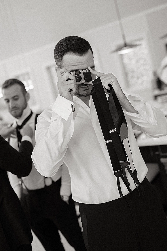 Groom getting ready adjusts suspenders and bow tie in a softly lit indoor room, with groomsmen nearby and a pendant light overhead