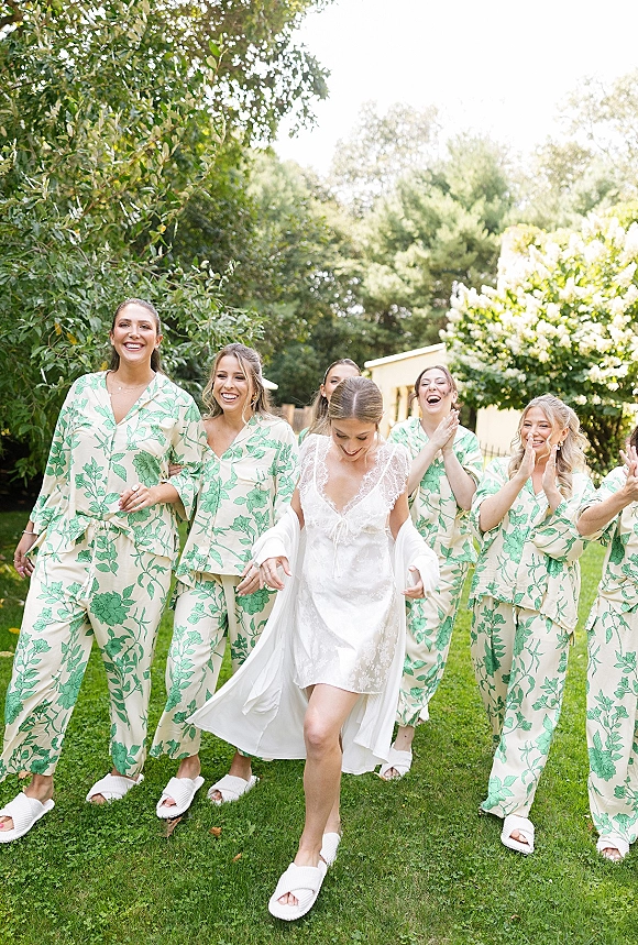 Bride with bridesmaids in matching pajamas cheer beside her white robe on a green lawn with garden trees and house exterior behind