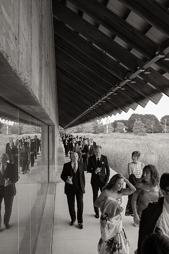 Wedding guest arrivals in formal attire walking with drink glasses under a covered walkway beside glass wall reflections at a modern venue exterior