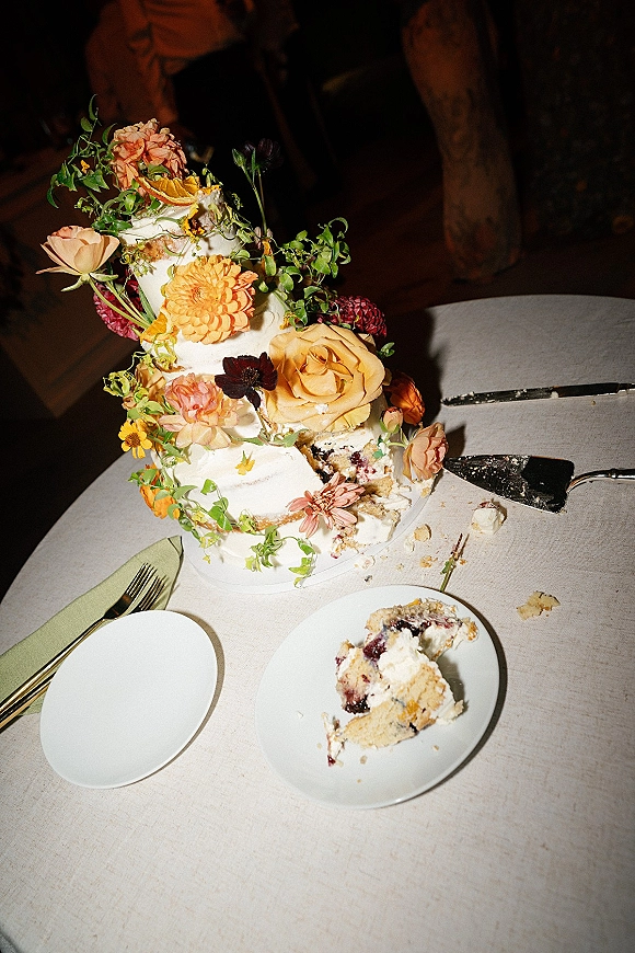 Wedding cake with floral wedding cake topper, greenery vines and citrus slices on a reception table in dim indoor lighting