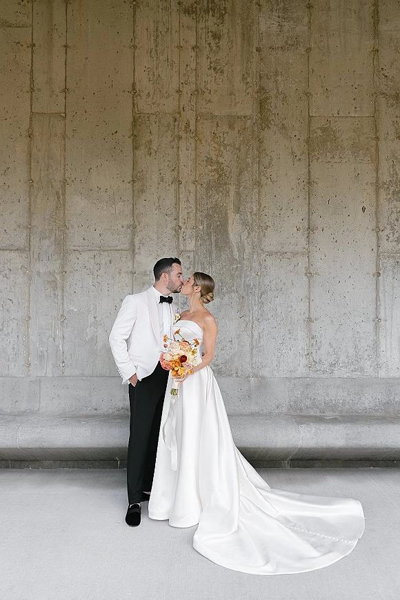 Wedding kiss portrait of bride and groom kissing, her strapless gown with long train and bouquet, against a concrete wall indoors