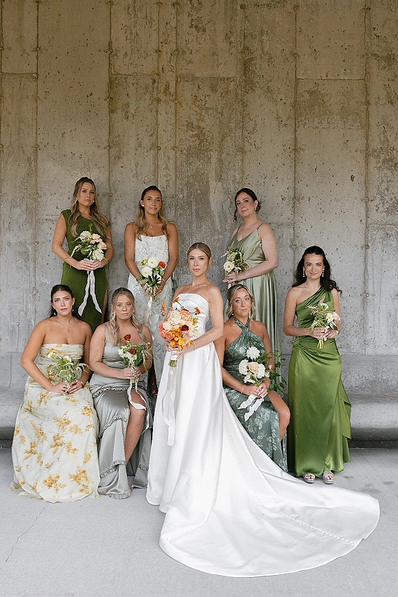 Bridesmaids portrait of bride with bridesmaids in green dresses, holding bouquets with ribbon ties against a modern concrete wall patio setting