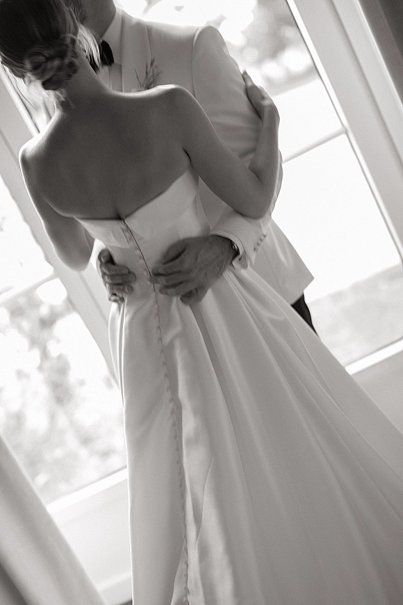 Wedding couple portrait sharing a wedding kiss moment, bride in strapless button-back gown and groom in bow tie by large windows