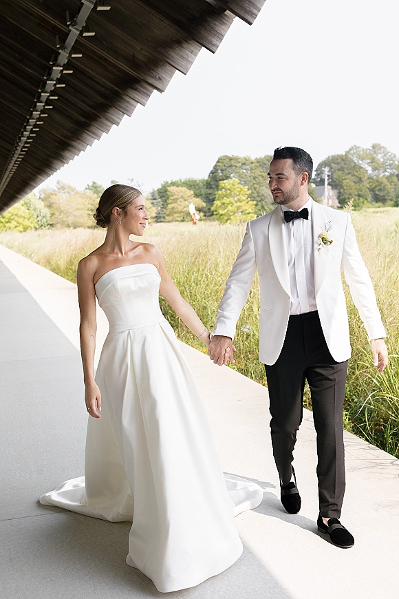 Couple portrait of bride and groom holding hands, her satin strapless gown with long train under a modern covered walkway by tall grass