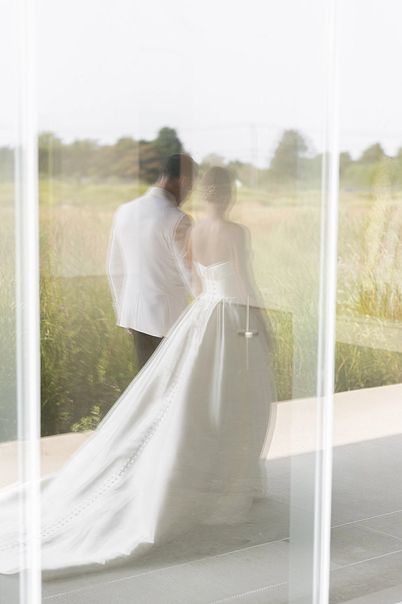 Couple portrait of bride and groom walking away, her strapless gown’s long train trailing by a glass window with field and trees beyond