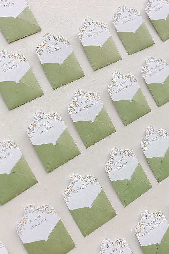 Wedding place cards in sage green envelopes with scalloped floral border cards and calligraphy names on a light neutral tabletop