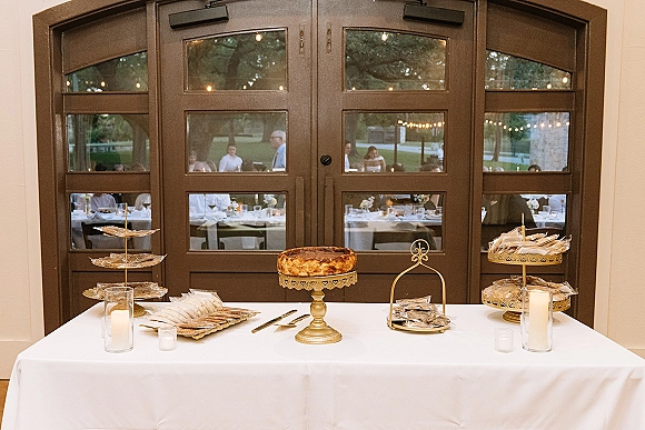 Dessert table with cheesecake and cookies on tiered stands, gold cake stand and glass cylinder candles by arched glass doors with string lights