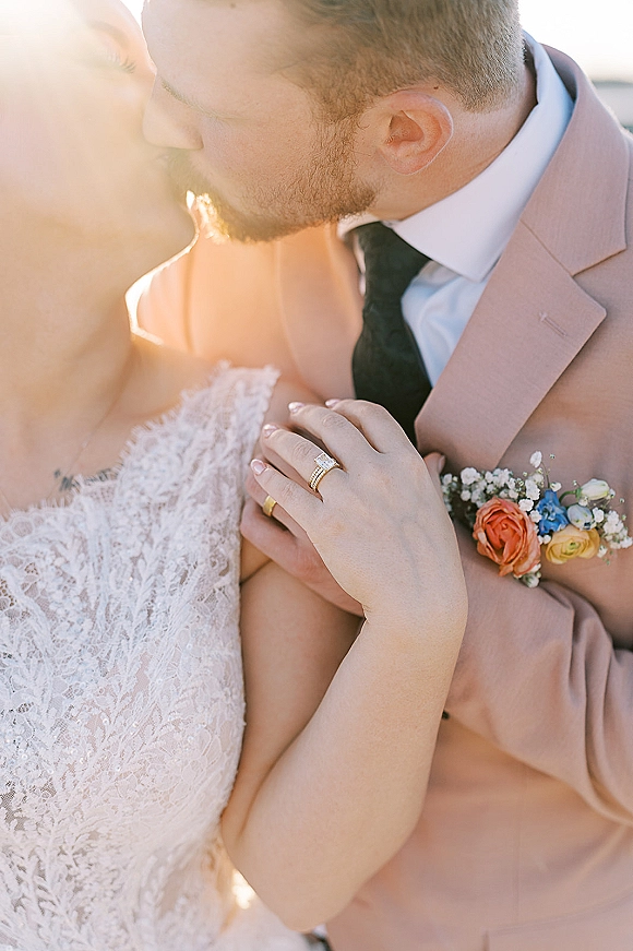 Wedding kiss close up as the groom kisses the bride’s forehead, her lace dress and rings catching a sun flare against the sky