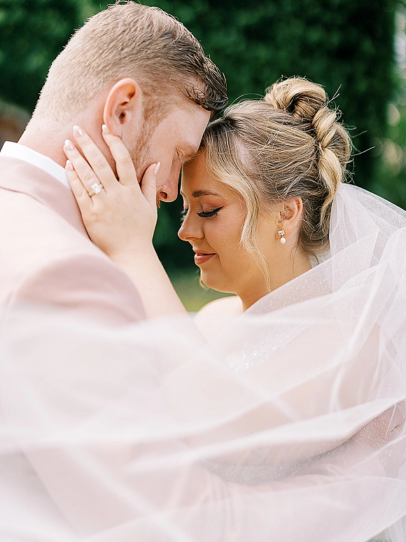 Couple portrait with bride and groom touching foreheads, her hand on his face showing engagement ring, veil accent, in garden trees backdrop