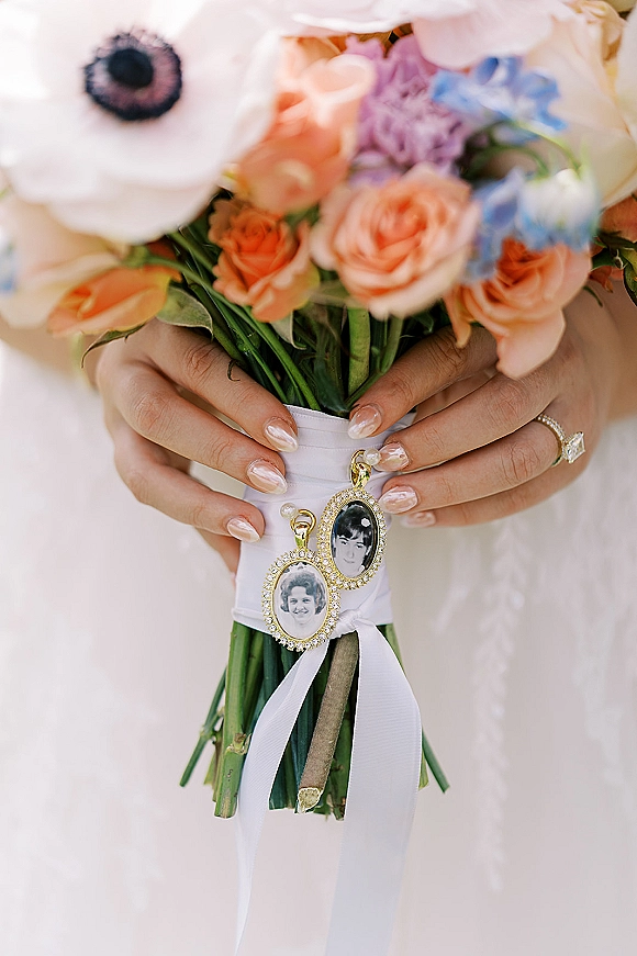 Wedding bouquet with peach roses and white anemone, wrapped in white ribbon, held over a white dress with locket charms and ring visible