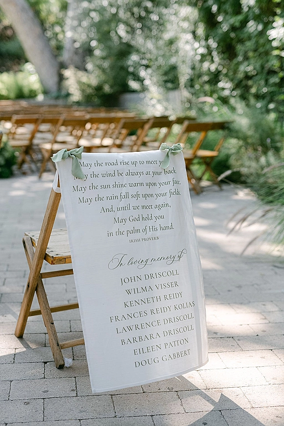Wedding memorial sign with in loving memory wedding sign calligraphy on a linen-draped wooden chair, sage green ribbon bows by a garden aisle
