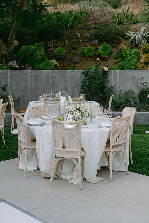 Reception tablescape with an outdoor reception table of white linens, cane-back chairs, pears, taper candles, and florals on a patio lawn