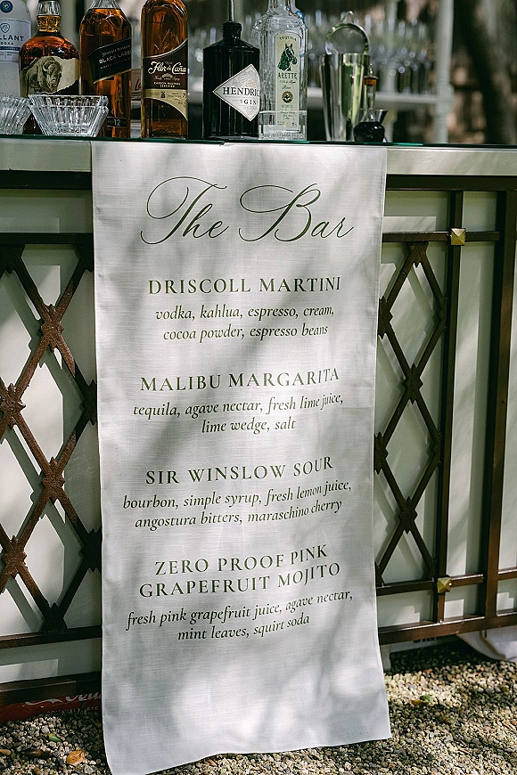 Wedding bar menu fabric sign with calligraphy lettering on a metal bar cart with liquor bottles, shaker, glassware, and mirror backdrop