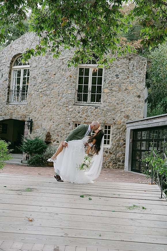Wedding kiss portrait of the groom lifting the bride in a dip kiss, her veil flowing, on a wooden deck before a stone building