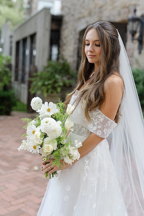Bridal portrait of a bride holding bouquet, looking down in a lace off-the-shoulder gown with cathedral veil on a brick walkway by a stone building