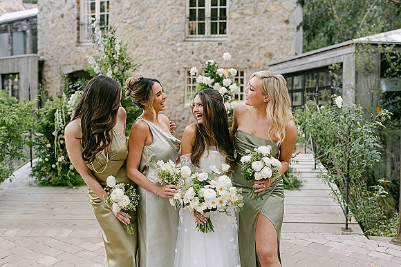 Bridesmaid group portrait with bride and bridesmaids holding bouquets in sage green dresses, posing by a stone building courtyard walkway