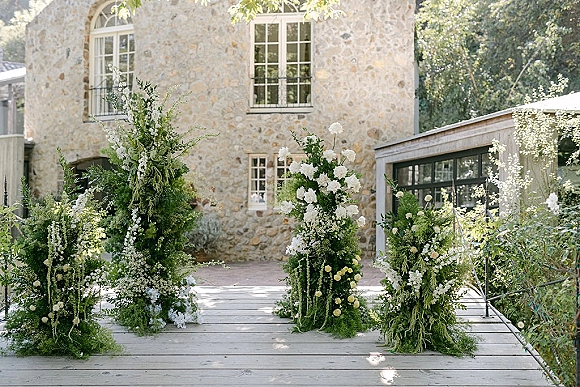Ceremony altar flowers in green and white, asymmetrical pillars with ferns and hanging greenery on a wooden platform in a stone courtyard