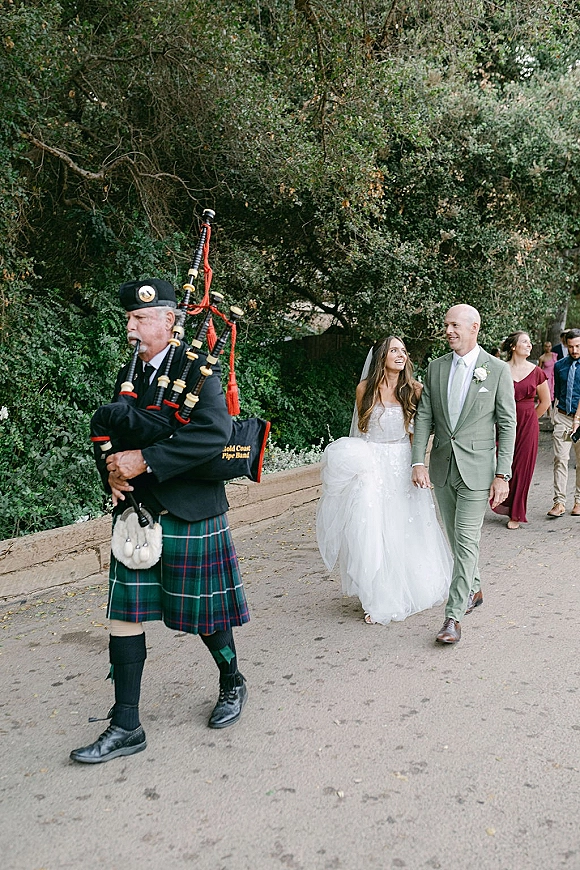 Wedding processional with bagpiper wedding entrance as bride in ball gown and veil walks beside groom in sage suit down tree-lined path
