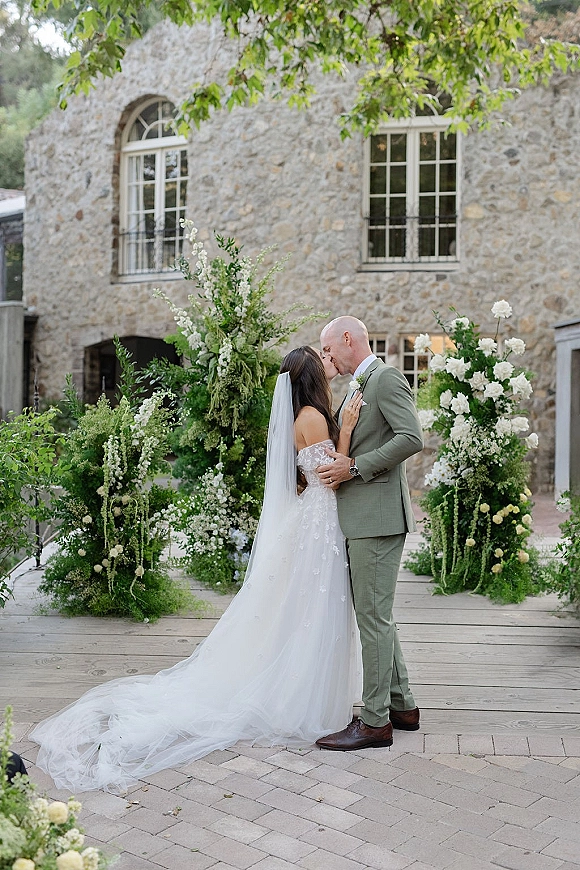 Wedding kiss as bride in lace gown and long veil embraces groom in sage suit before white florals in a stone courtyard venue