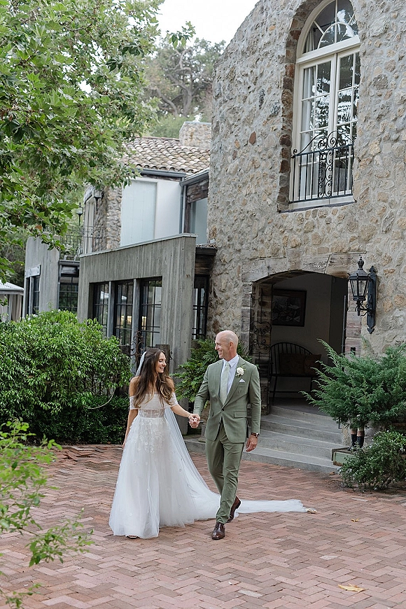 Couple portrait of bride and groom walking hand in hand, her long veil and dress train flowing outside a stone building courtyard with arched window