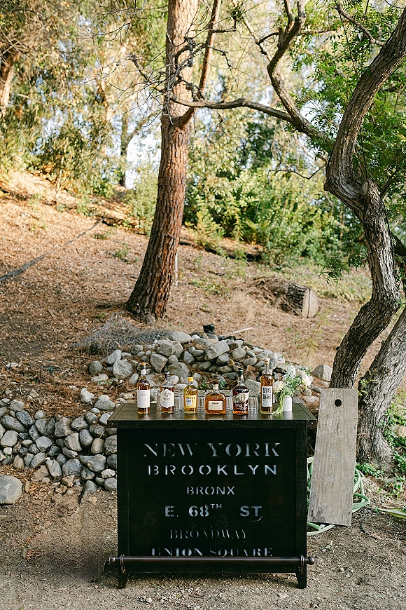 Wedding bar setup with liquor bottles on a black bar cabinet and printed sign, accented by a small floral arrangement in a wooded setting
