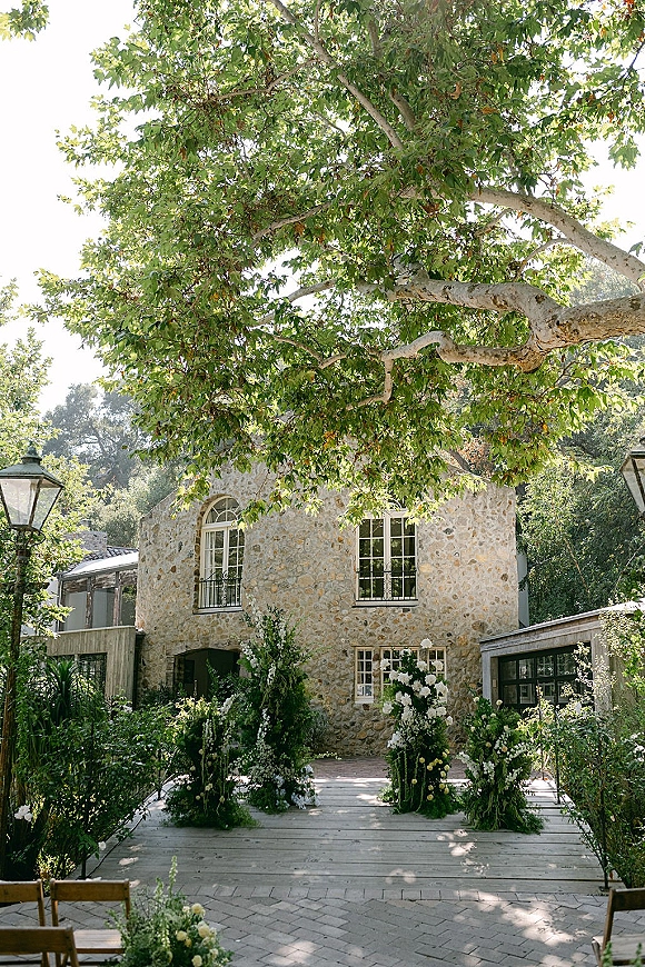 Ceremony setup with an outdoor ceremony aisle lined in white-green florals and wooden chairs, set before a stone villa courtyard facade