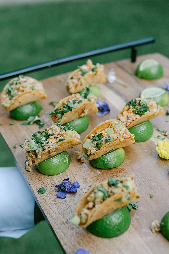 Wedding cocktail appetizers and wedding hors d'oeuvres of mini tacos with lime halves and herb garnish on a wooden board on a green lawn