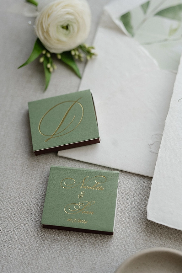 Wedding invitation suite with sage green wedding invitations in boxes, gold foil monogram, white envelope, and ranunculus boutonniere on neutral tabletop