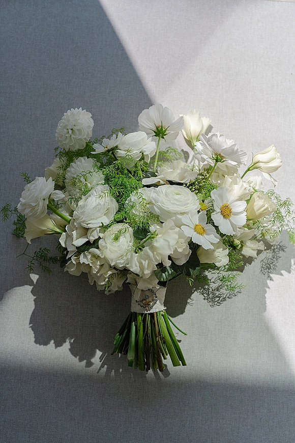 Bridal bouquet, a white wedding bouquet of white blooms and greenery with wrapped stems, resting on gray fabric in soft window light shadows