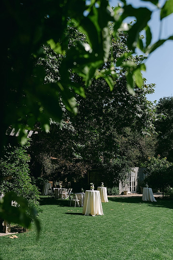 Cocktail hour setup with outdoor cocktail tables draped in white linens, bud vases of white flowers, and folding chairs on a sunlit garden lawn near a shed