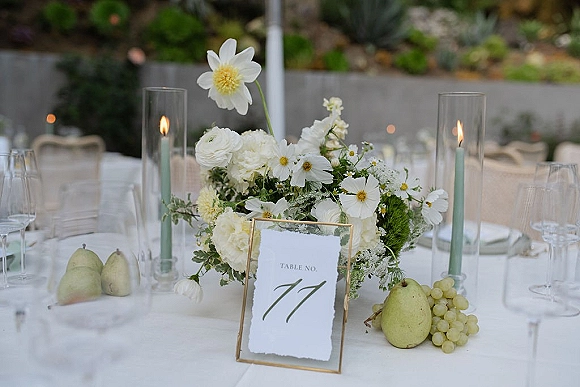 Reception tablescape with a wedding table centerpiece of white flowers and greenery, green taper candles, fruit, and gold table number in a garden setting